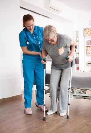 Old woman in nursing home walking with crutches with help from female nurse.